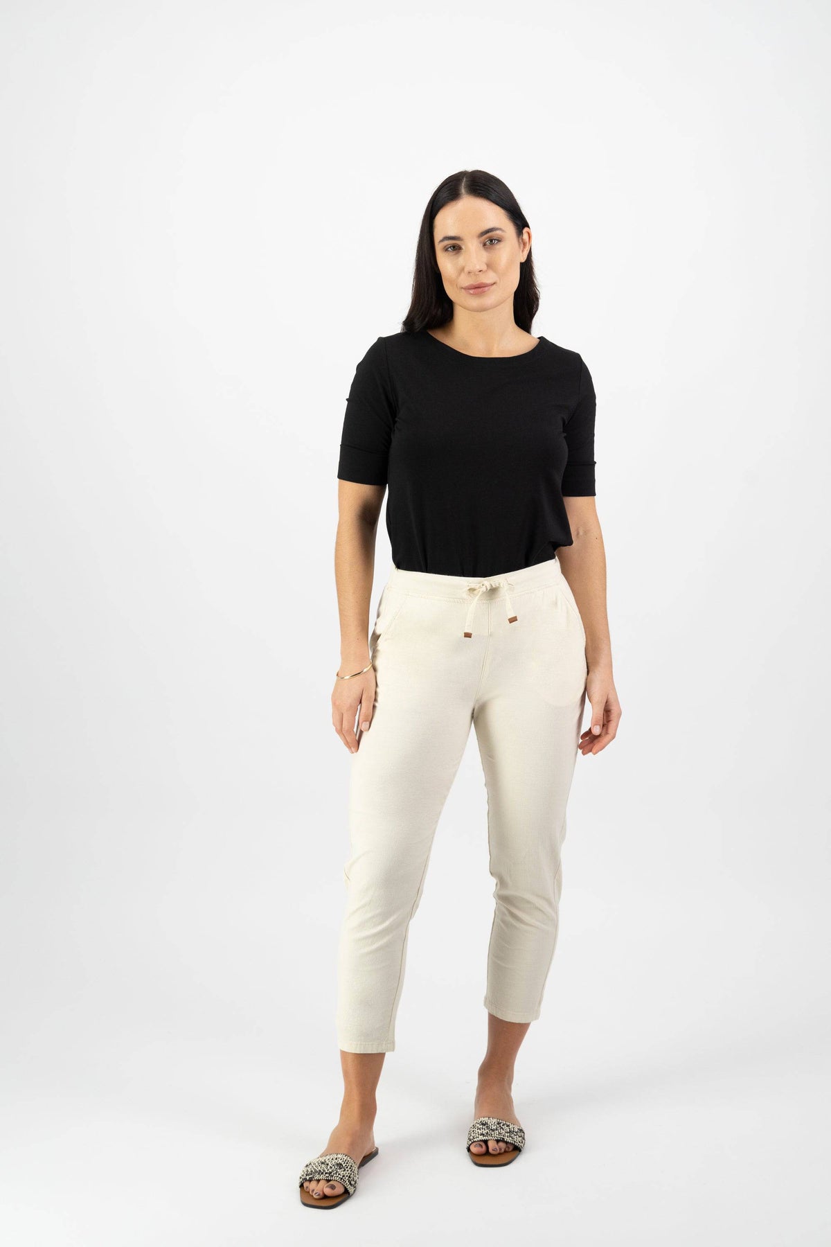 A woman with long dark hair stands against a white background, wearing a black short-sleeve top and Vassalli Denver 7/8 Pant in Ecru, featuring an elastic waist and skinny leg. She has one hand by her side and the other in her pocket.