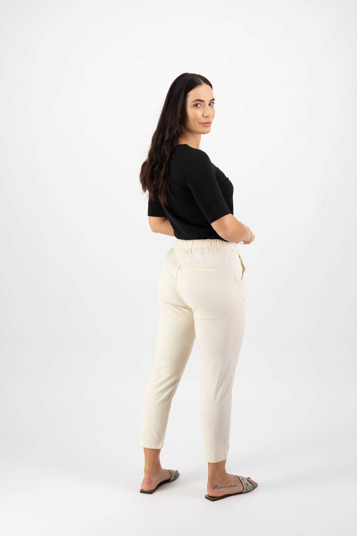 A woman with long dark hair stands sideways, looking over her shoulder. She wears a black top, Vassalli Denver 7/8 Pant in Ecru with an elastic waist and skinny leg, and slip-on shoes, posing against a plain white background.