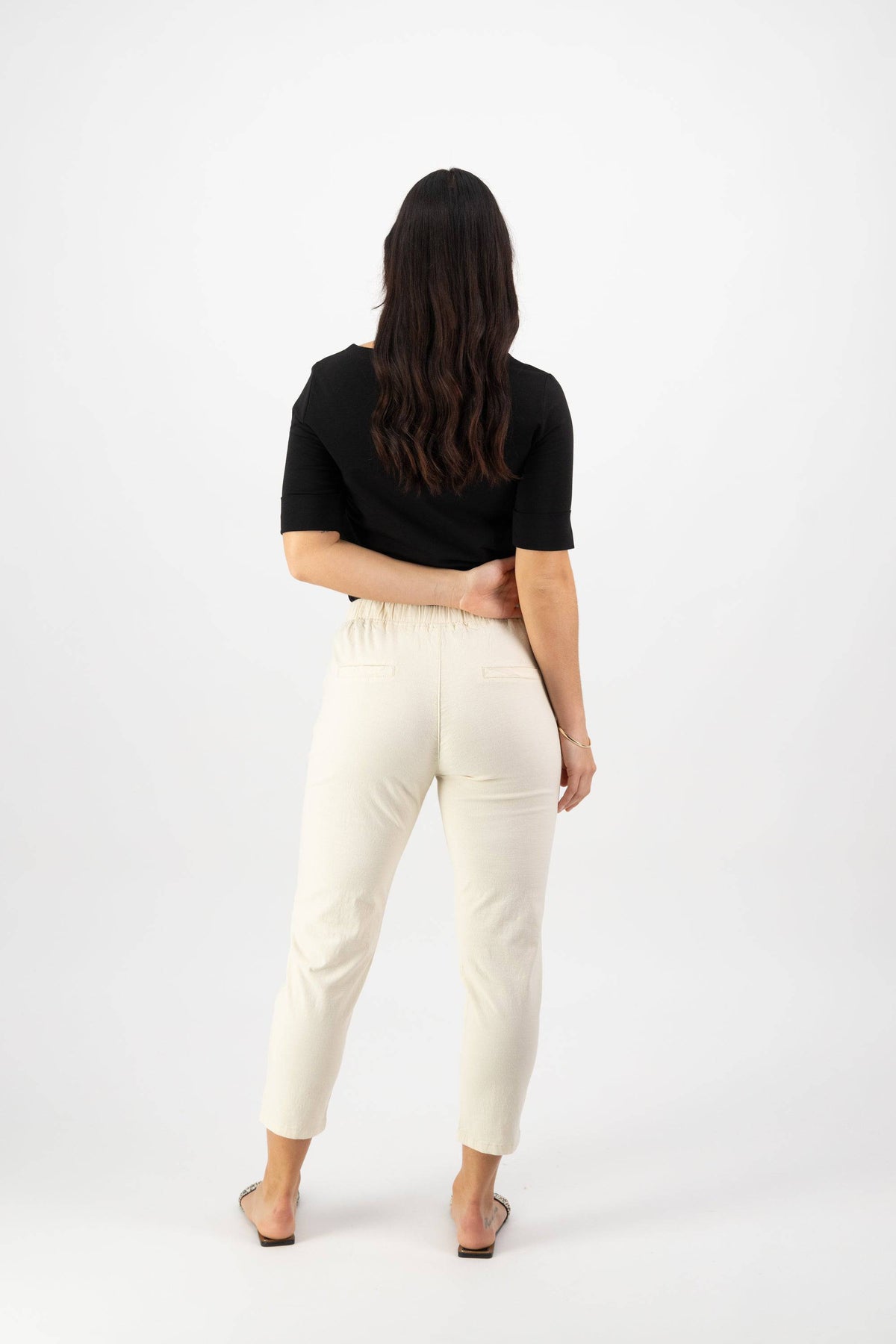 A woman with long brown hair in a black top and Vassalli Denver 7/8 Pant - Ecru (skinny fit) stands facing away from the camera, one arm bent behind her back, against a plain white background.