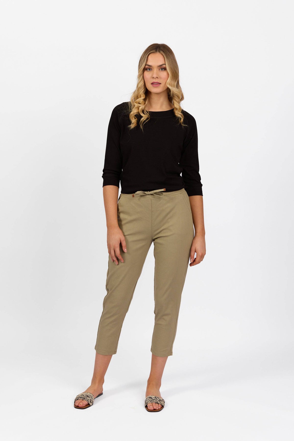 A woman with long blonde hair wears a black long-sleeve top and Vassalli Denver 7/8 Pant in Sage, styled with sandals. She stands against a plain white background, looking at the camera with a neutral expression.