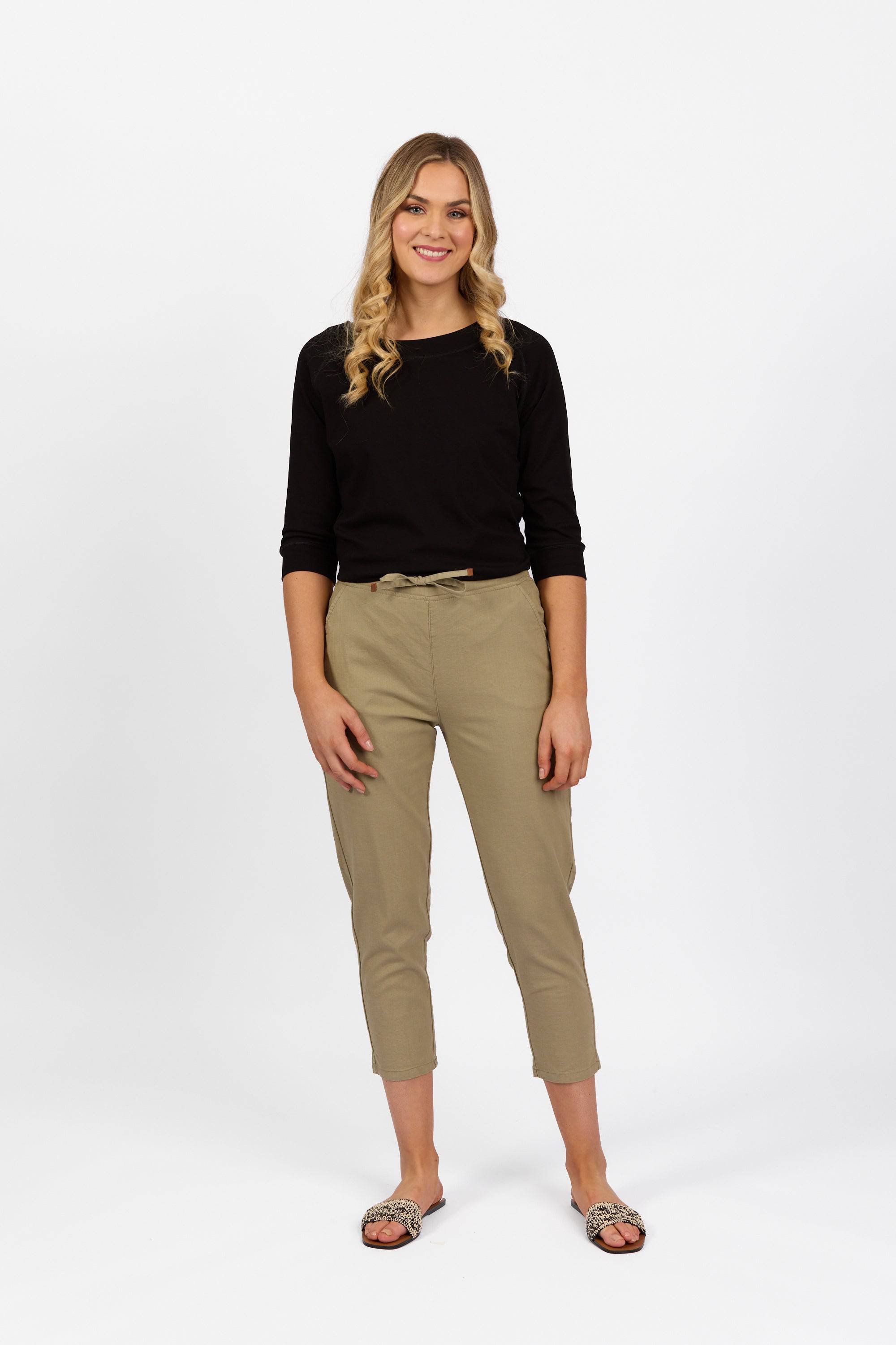 A woman with long wavy hair wearing a black long-sleeve top, Vassalli Denver 7/8 Pant in Sage, and sandals stands smiling against a plain white background.