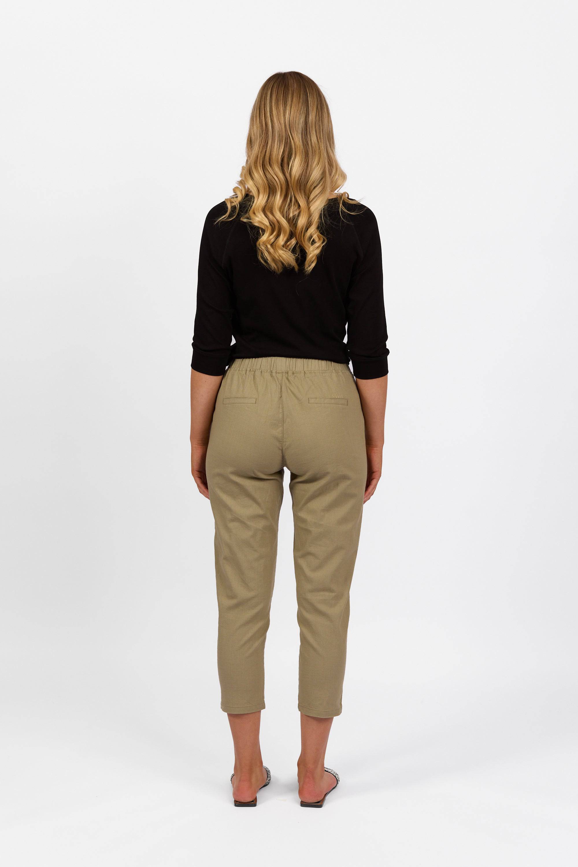 A woman with long wavy blonde hair, barefoot and facing away from the camera, wears a black long-sleeve top and Vassalli Denver 7/8 Pant in Sage with an elastic waist, set against a plain white background.