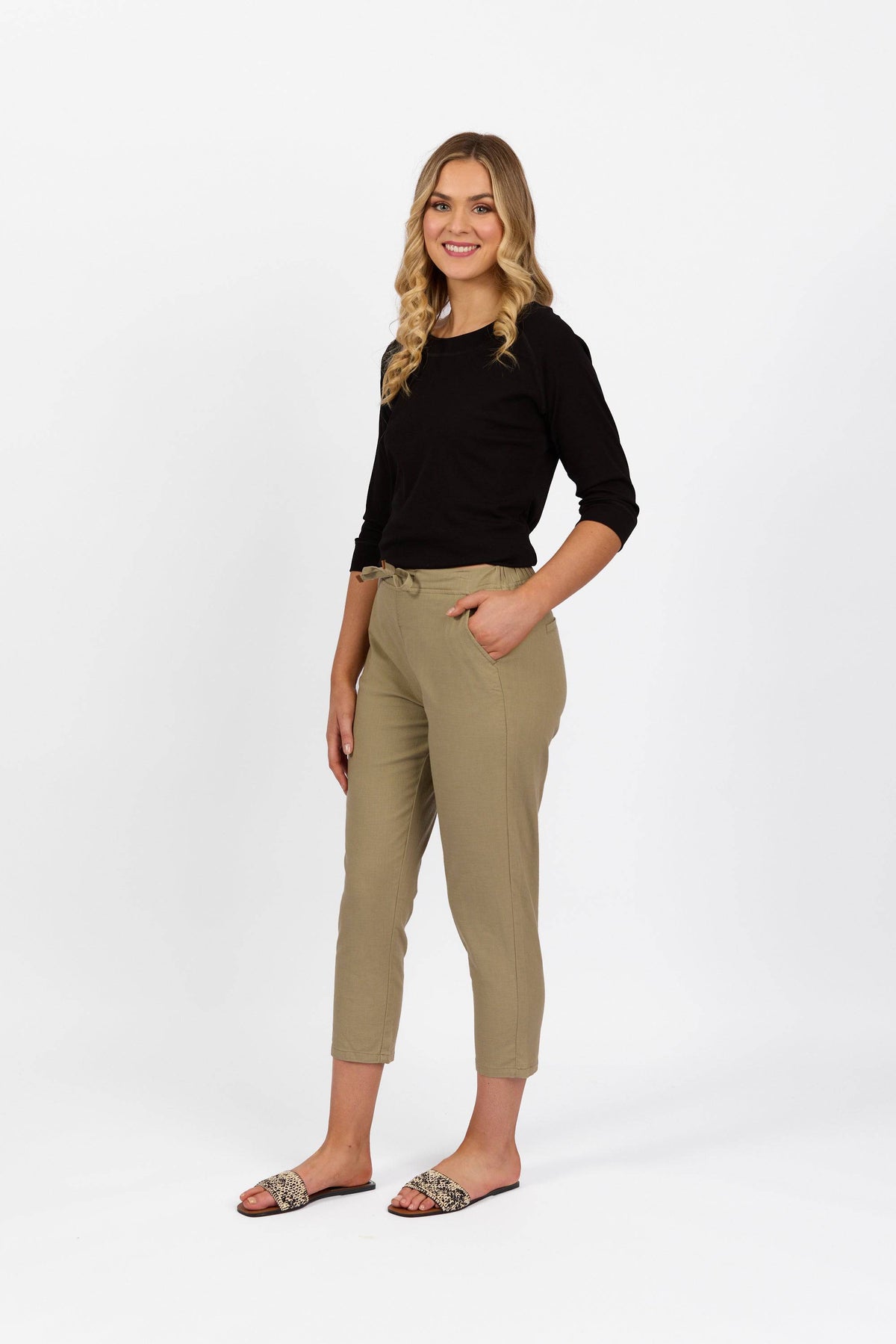 A woman with long blonde hair in a black top and Vassalli Denver 7/8 Pant - Sage stands smiling, one hand in her pocket, against a white background. She completes her look with patterned slip-on shoes.