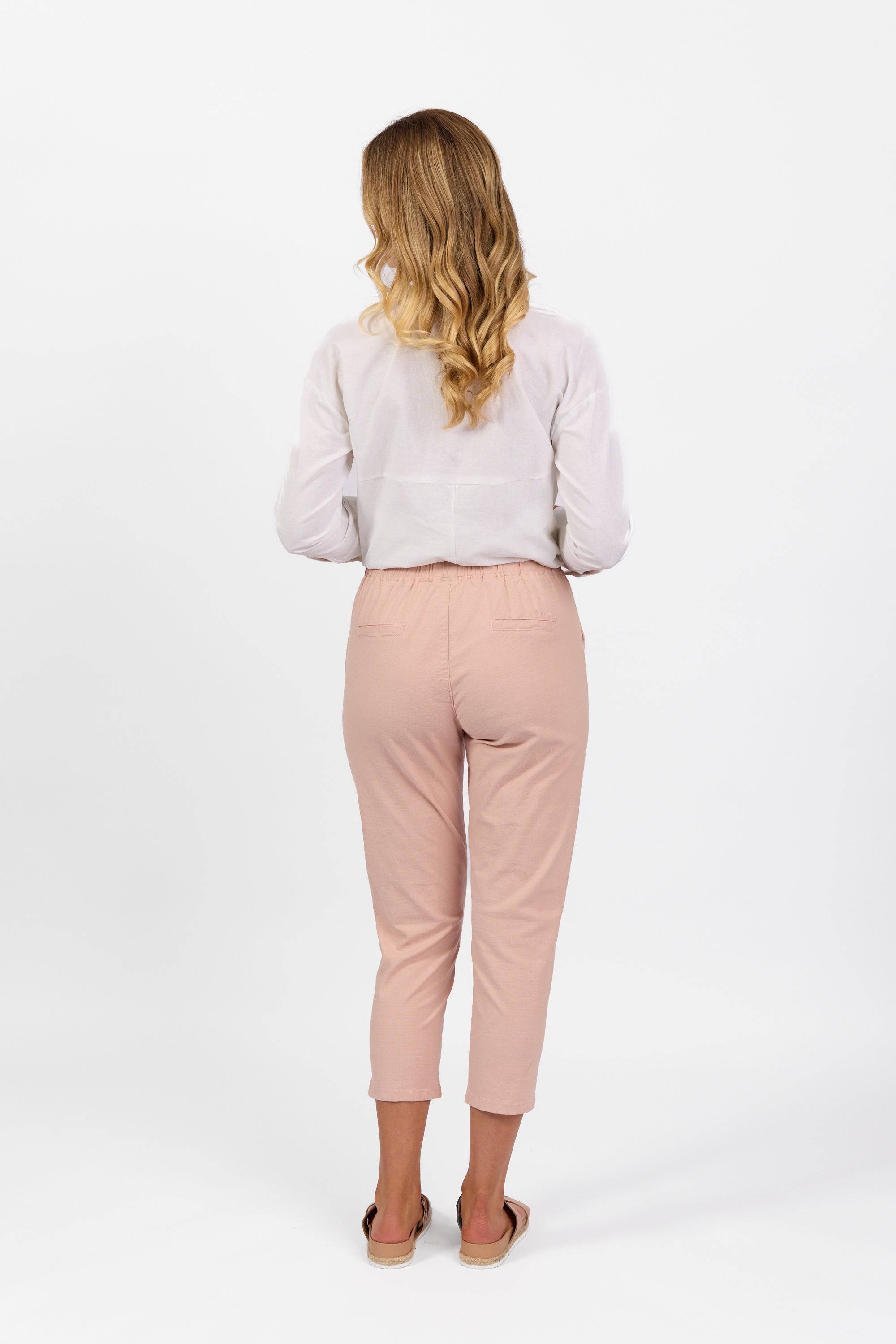 A woman with long, wavy blonde hair, seen from behind, wears a white blouse and Vassalli Denver 7/8 Pant in Soft Pink with an elastic waist, paired with beige slip-on shoes against a plain white background.