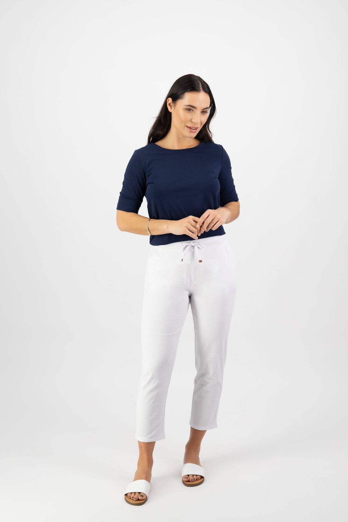 A woman with long dark hair stands against a white background, smiling slightly as she touches the waistband of her Vassalli Denver 7/8 Pant in White, styled with a navy top.