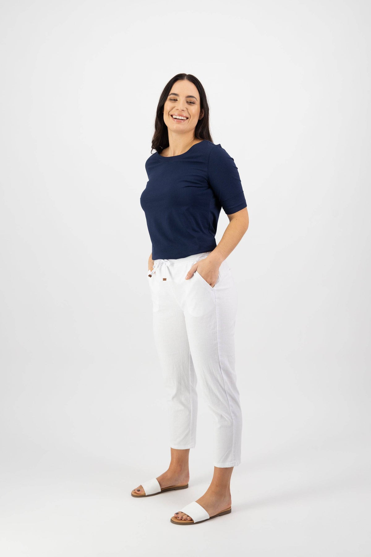 A woman smiles with her hands in the pockets of her Vassalli Denver 7/8 Pant in white, pairing them with a navy blue top and white slide sandals, set against a plain white background.