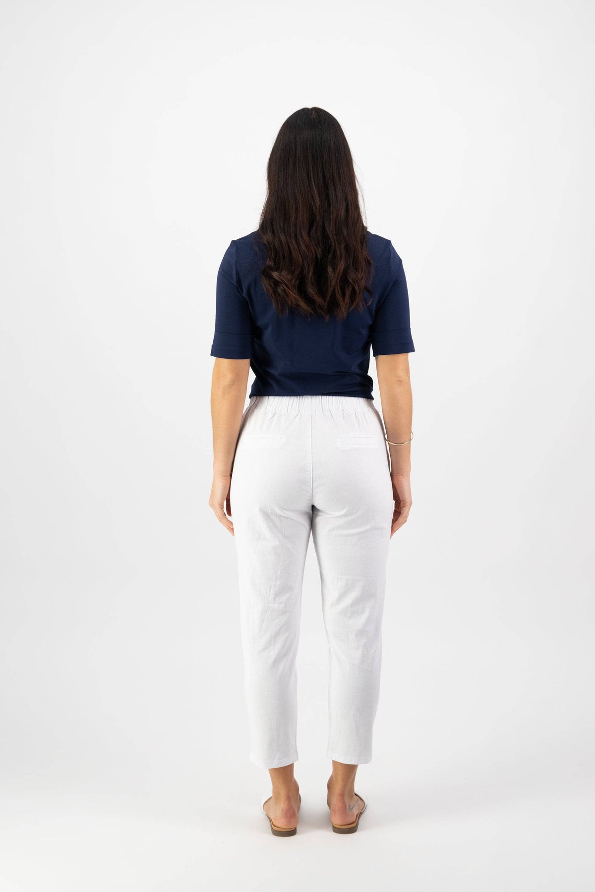 A person with long dark hair faces away, wearing a navy blue short-sleeve top, Vassalli Denver 7/8 Pant in white (cotton blend, elastic waistband), and light-colored sandals against a plain white background.