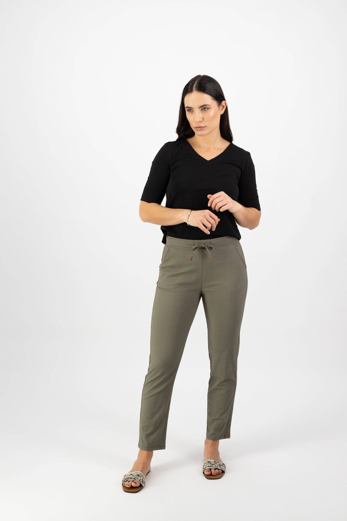 A woman with long dark hair stands against a white background, wearing a black short-sleeve top, Vassalli Leonie 7/8 Pant in Army, and leopard print shoes. She looks to the side with a neutral expression.