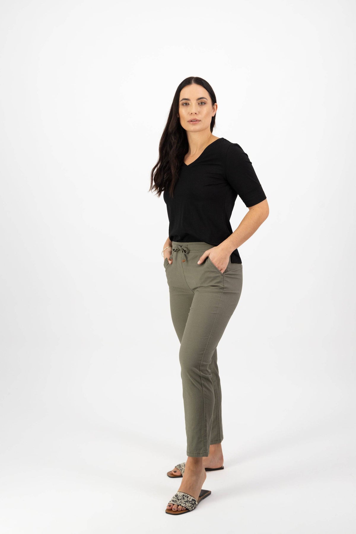 A woman with long dark hair, in a black short-sleeve shirt and leopard print sandals, stands with her hands in her pockets wearing Vassalli Leonie 7/8 Pant - Army against a plain white background.