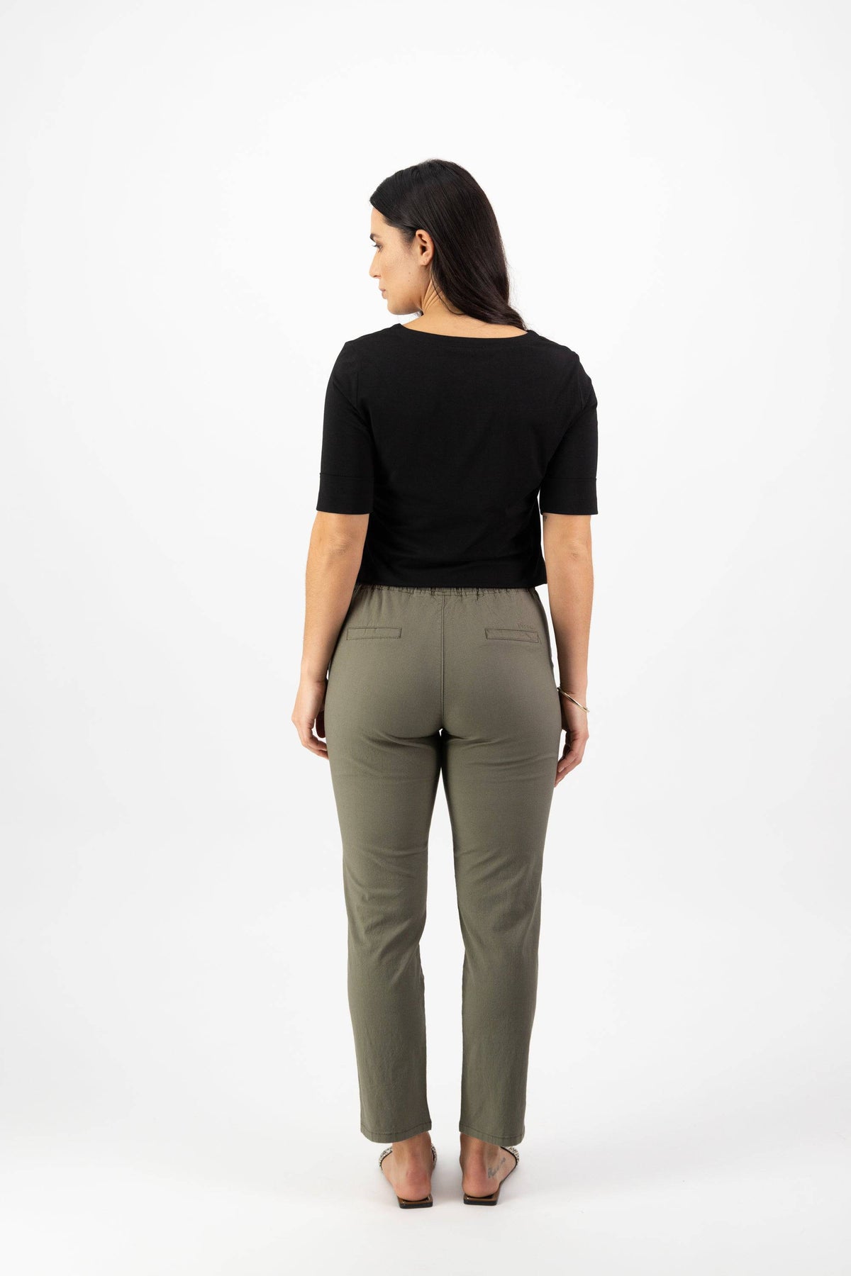 A woman with long dark hair faces away, wearing a black short-sleeve top and Vassalli Leonie 7/8 Pant - Army, crafted from stretch fabric, paired with sandals against a plain white background.