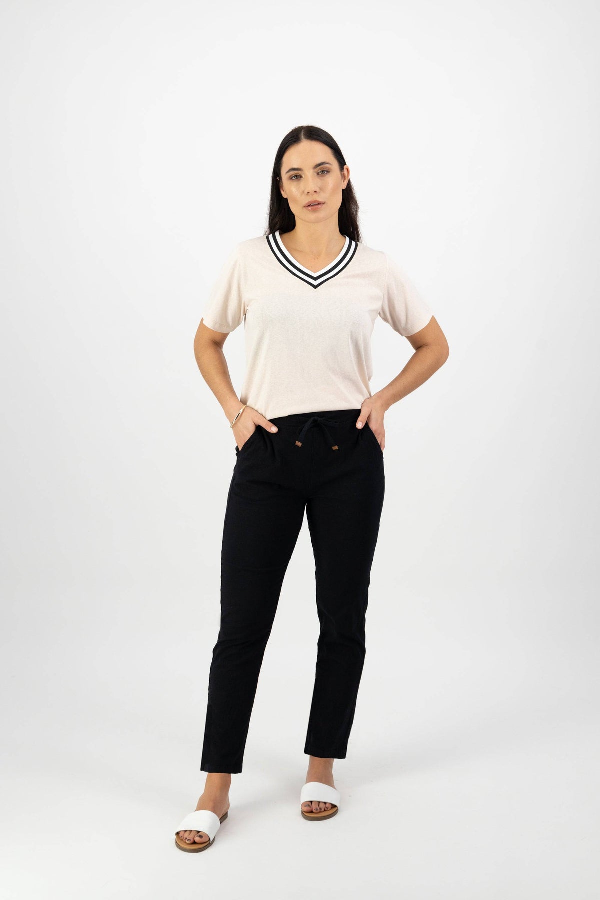 A woman with a neutral expression stands against a white background, wearing Vassalli's Leonie 7/8 Pant in black, a light V-neck short-sleeve top, and white slide sandals, with her hands in her pockets.