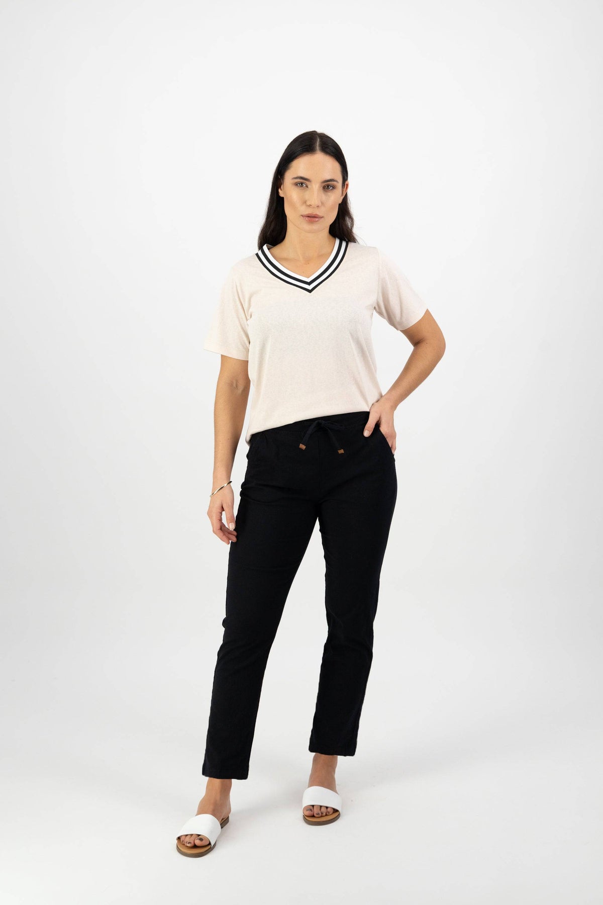 A woman with long dark hair stands against a white background wearing Vassalli's Leonie 7/8 Pant in black, styled with a light short-sleeve V-neck top with black trim, stretch fabric for comfort, white sandals, and a neutral expression.