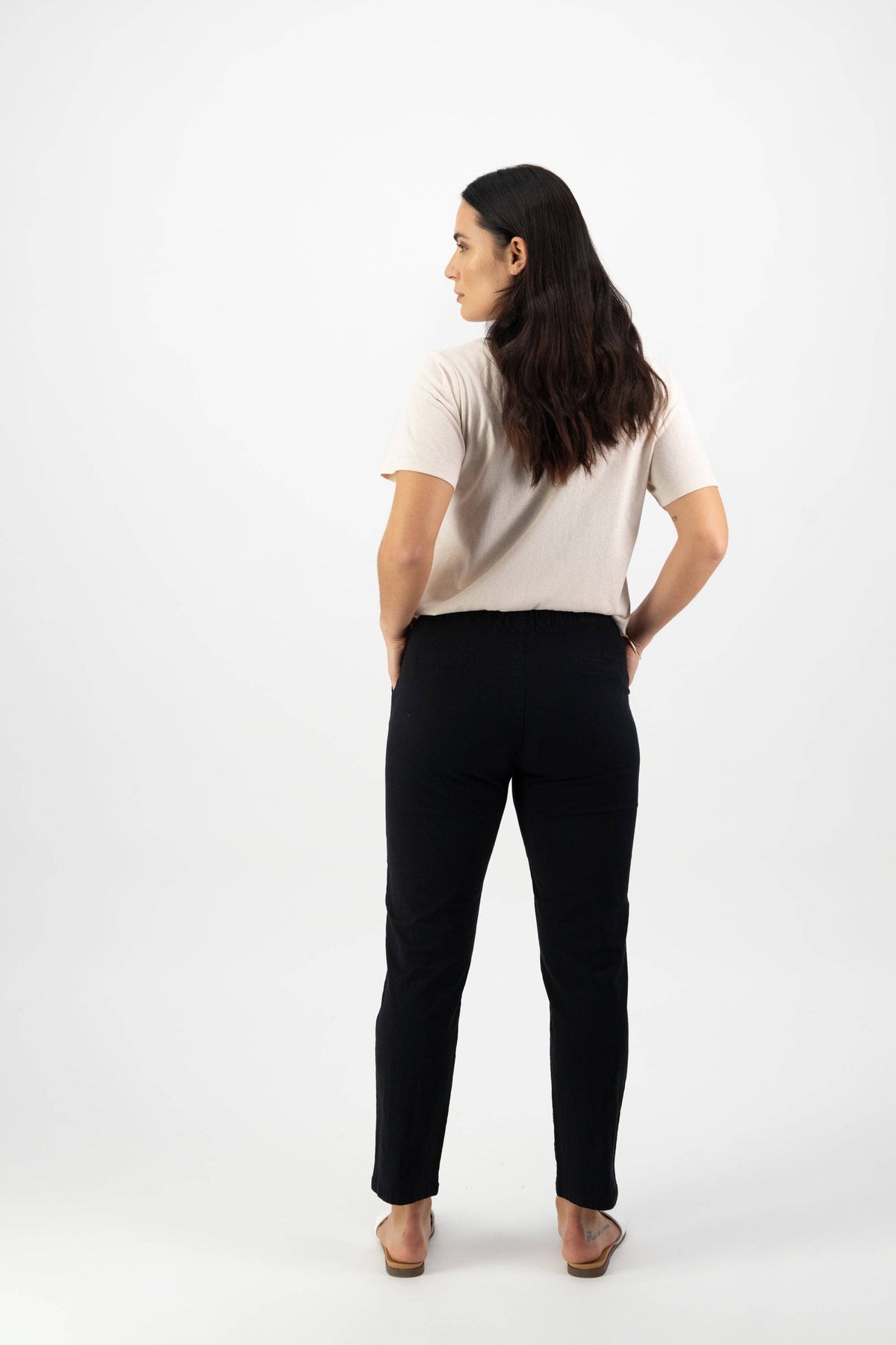 A woman with long dark hair stands facing away, hands in her pockets, wearing a beige top, Vassalli Leonie 7/8 Pant in Black, and sandals against a plain white background.