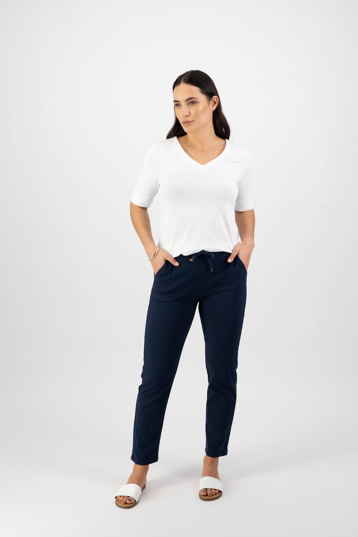 A woman stands against a white background wearing a white V-neck top, Vassalli Leonie 7/8 Pant in Ink, and white slide sandals. Her hands are in her pockets and she has a neutral expression.