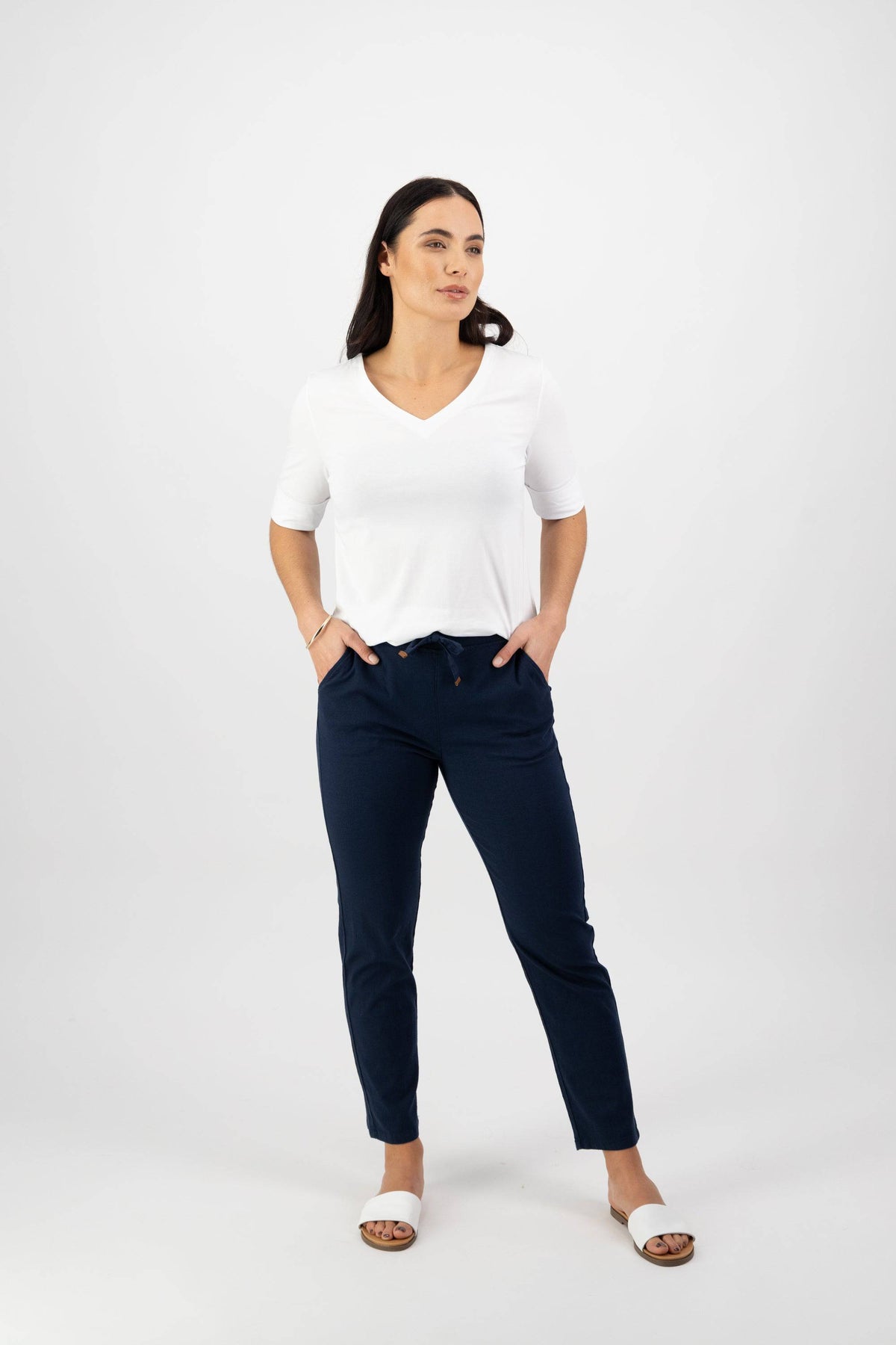 A woman with long dark hair stands confidently against a white background, wearing a white V-neck top, Vassalli's Leonie 7/8 Pant in Ink, and white slide sandals. Her hands are in her pockets as she gazes to the side.