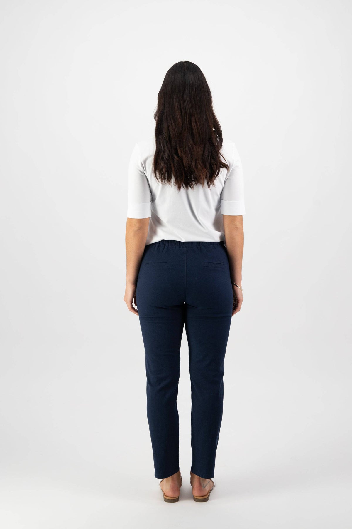 A person with long dark hair stands facing away, wearing a white short-sleeve top, Vassalli Leonie 7/8 Pant in Ink, and sandals against a plain white background.