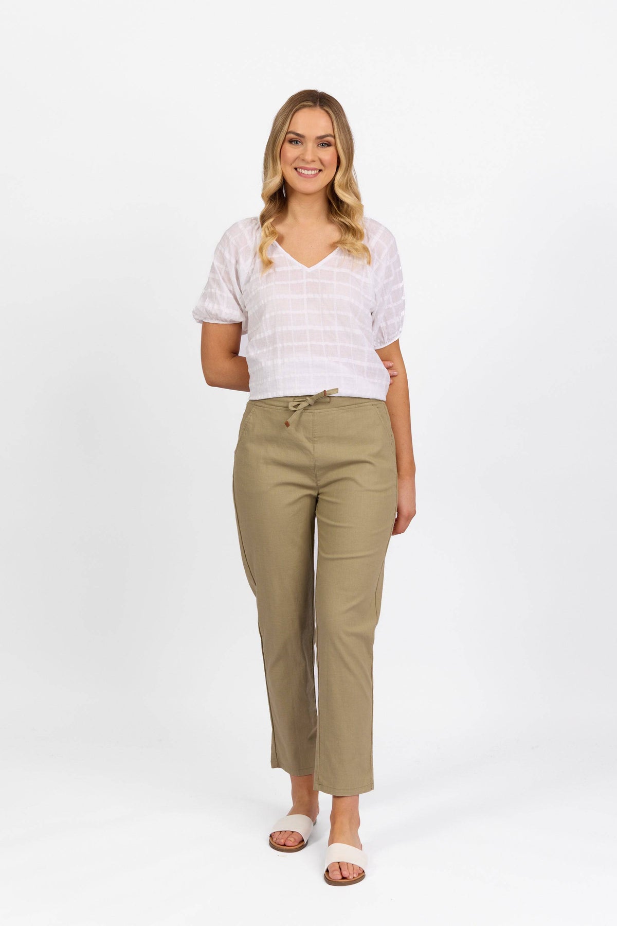 A woman stands and smiles, wearing a white short-sleeve top paired with the Vassalli Leonie Pant in Sage—crafted from stretch fabric with a slim-leg, elastic-waist design—and sandals, set against a plain white background.