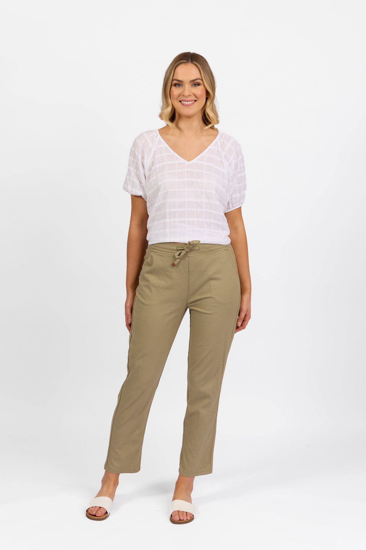A woman stands smiling, wearing a white short-sleeve blouse paired with the Vassalli Leonie Pant in sage, which features a simple tie belt and is crafted from comfortable stretch fabric. She completes her look with flat sandals, all set against a plain white background.