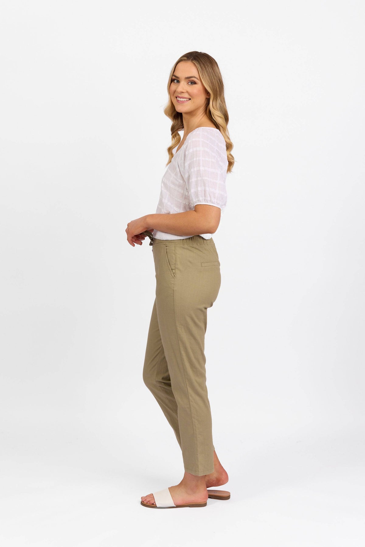 A woman stands sideways, smiling, wearing a white short-sleeve blouse and the Vassalli Leonie Pant in Sage, crafted from stretch fabric for comfort, paired with white slide sandals against a plain white background.