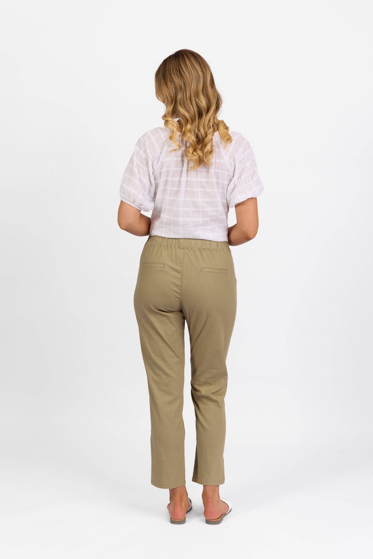 A person with long, wavy blonde hair stands facing away, wearing a short-sleeve white shirt and the Vassalli Leonie Pant in Sage, crafted from stretch fabric, paired with sandals against a plain white background.