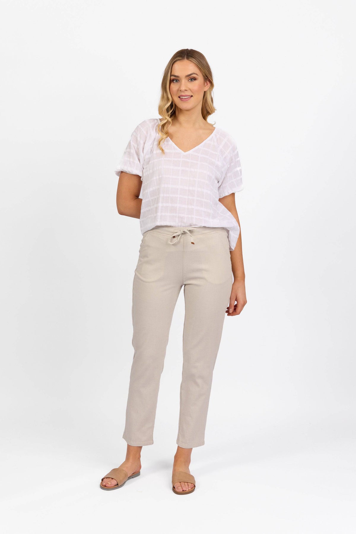 A woman stands against a white background wearing a white short-sleeve V-neck top and Vassalli Leonie 7/8 Pant in Stone, featuring a drawstring waist and slim leg stretch fit. She pairs them with tan slide sandals and has one arm behind her back.