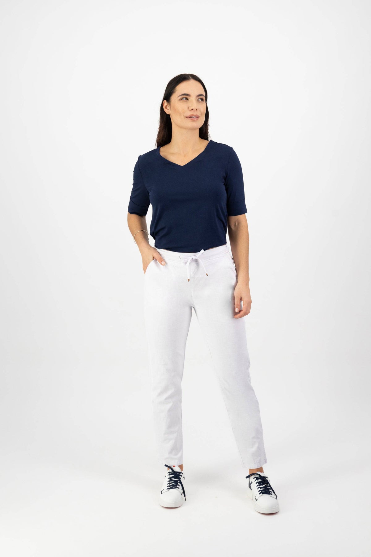 A woman stands against a white background, wearing the Vassalli Leonie 7/8 Pant in White with a navy blue short-sleeve top and black-and-white sneakers, looking slightly to the side with a neutral expression.