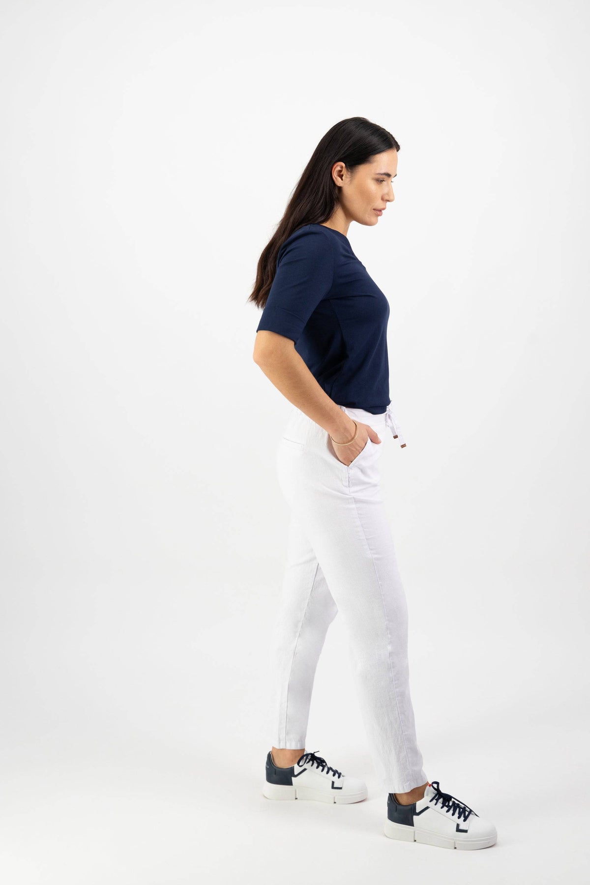 A woman with long brown hair, wearing a navy blue top and Vassalli Leonie 7/8 Pant in white with an elastic waistband, stands in profile with hands in pockets against a plain white background.