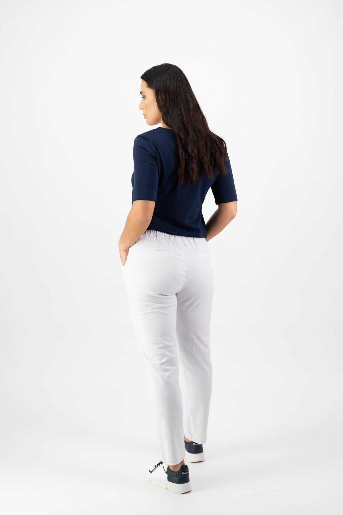 A woman with long brown hair, wearing a navy top and Vassalli's Leonie 7/8 Pant in white with an elastic waistband, stands facing away in black sneakers, looking to the side against a plain white background.