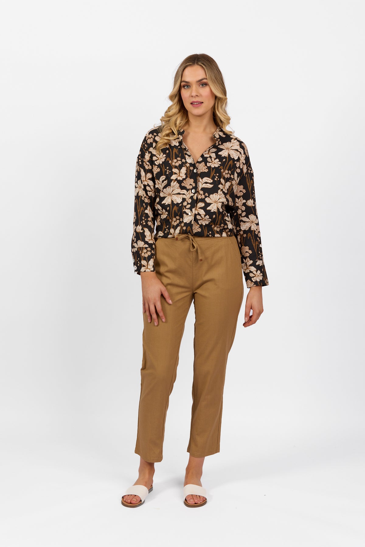 A woman with blonde wavy hair poses confidently against a white background, wearing the Vassalli Leonie Pant in Caramel, styled with a black floral blouse and beige sandals.