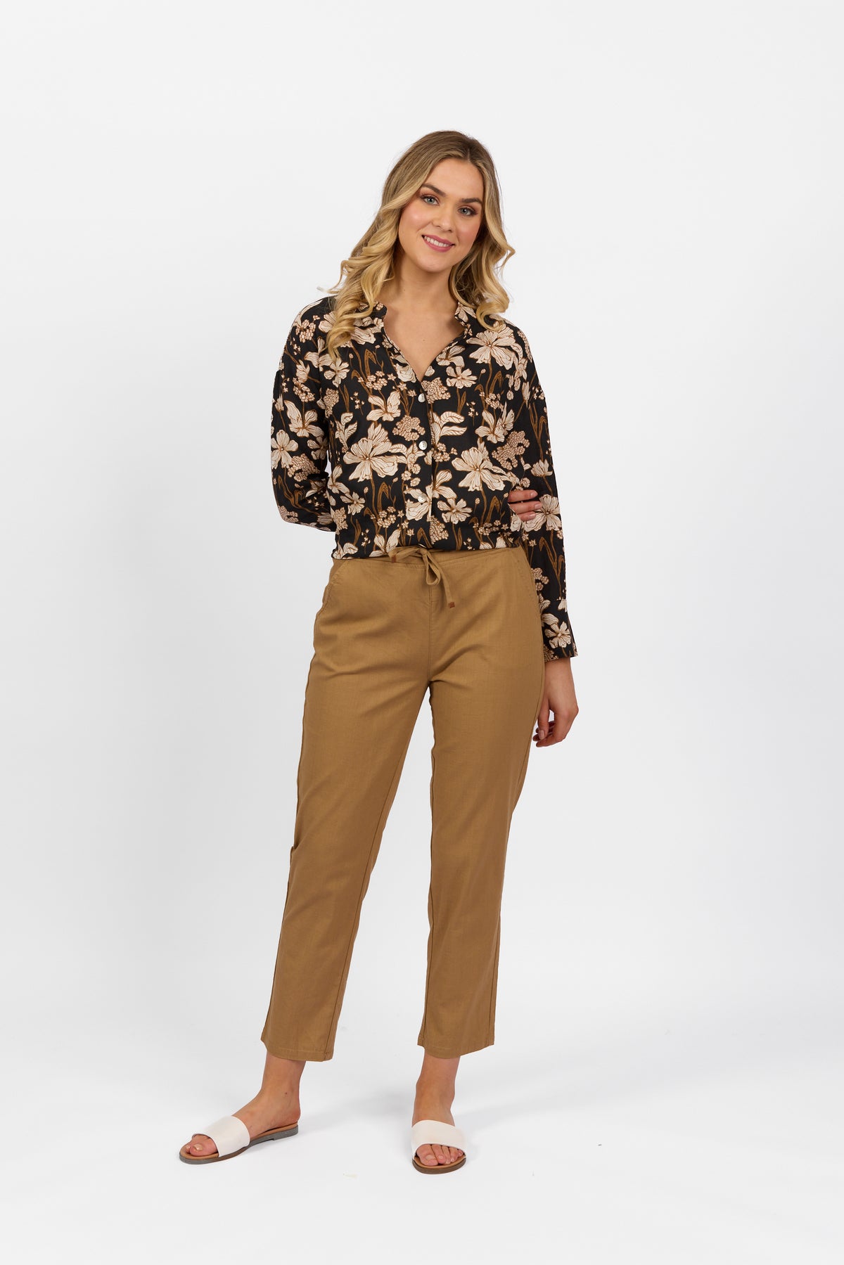 A woman with long, wavy blonde hair smiles with one hand in her pocket, wearing the Vassalli Leonie Pant in Caramel, a black floral long-sleeve blouse, and white slide sandals against a white background.