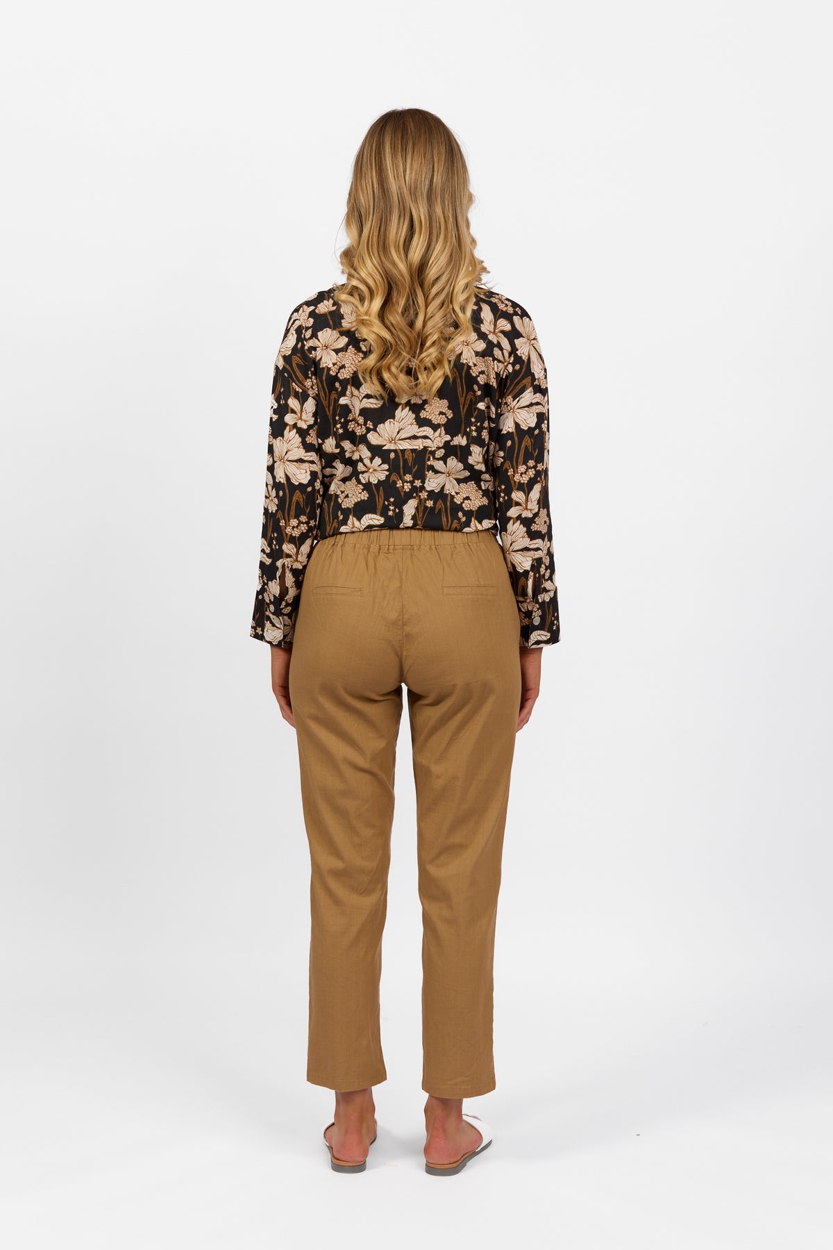 A person with long, wavy blonde hair faces away, wearing a black floral top, Vassalli Leonie Pant in Caramel with an elastic waist and slim leg, and sandals against a plain white background.