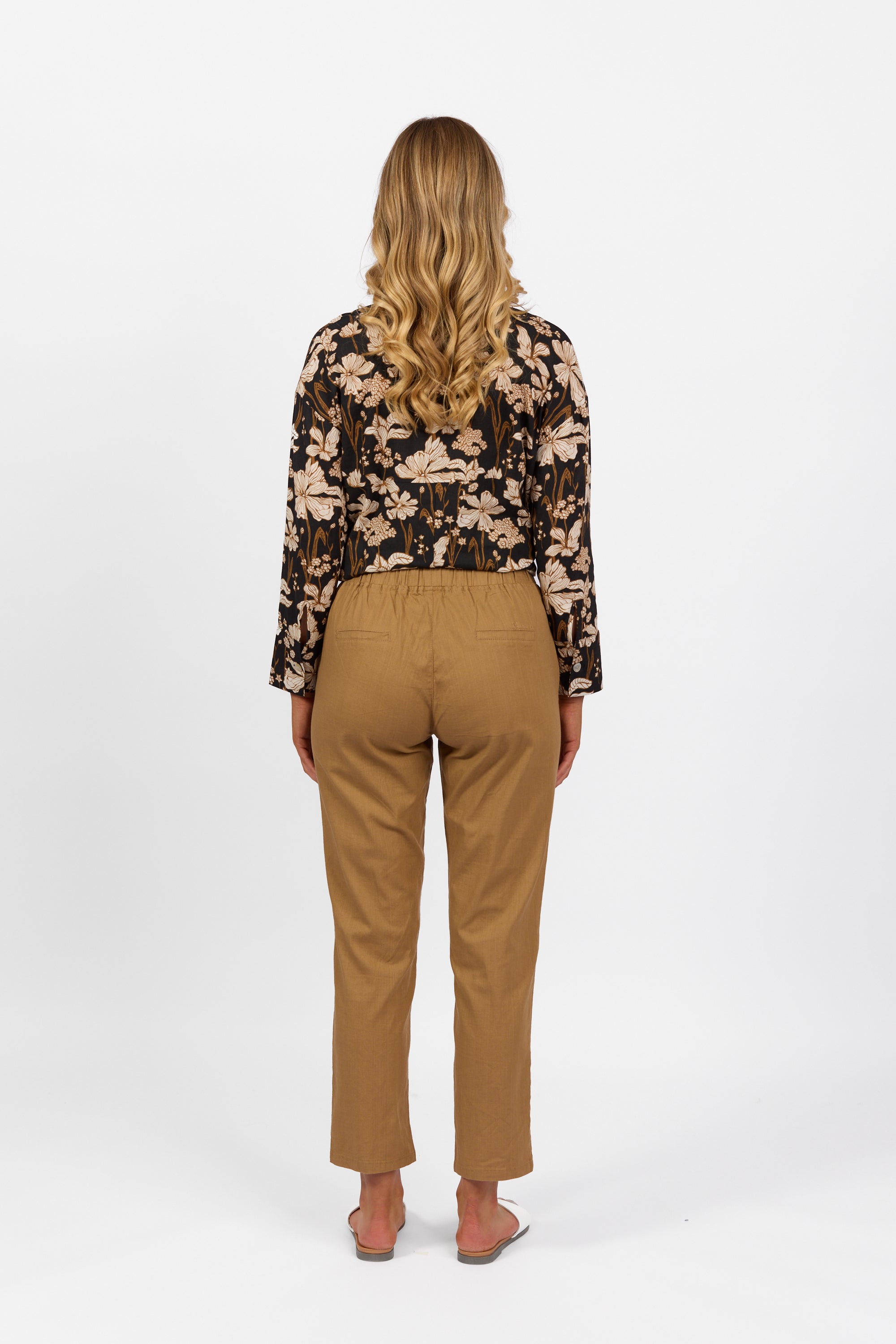 A person with long, wavy blonde hair faces away, wearing a black floral top, Vassalli Leonie Pant in Caramel with an elastic waist and slim leg, and sandals against a plain white background.