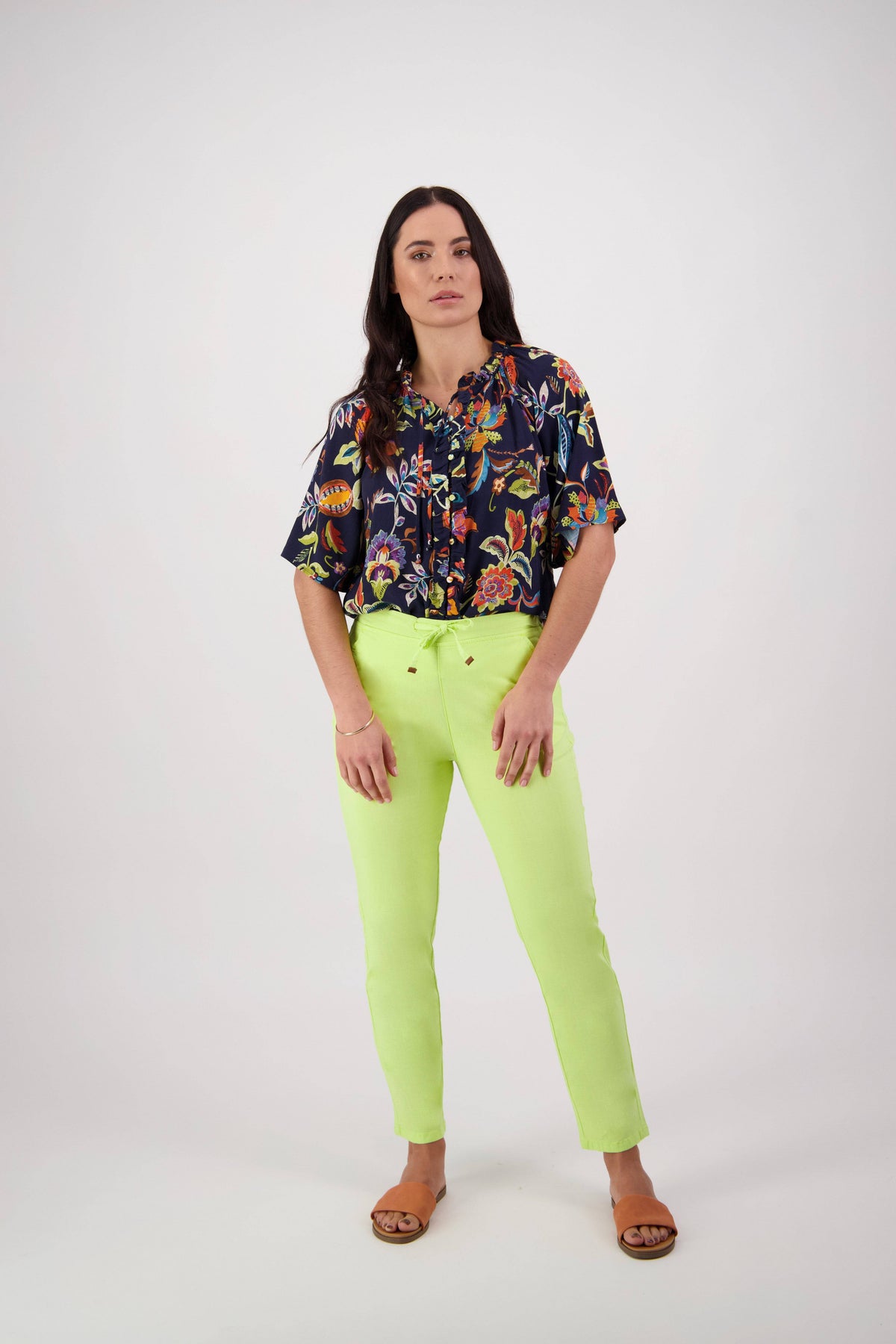 A woman with long dark hair stands against a white background, facing forward with a neutral expression. She is wearing a dark floral blouse, brown sandals, and the Vassalli Leonie Pant in Lime—an ankle-grazer style featuring a slim leg and an elastic waist.