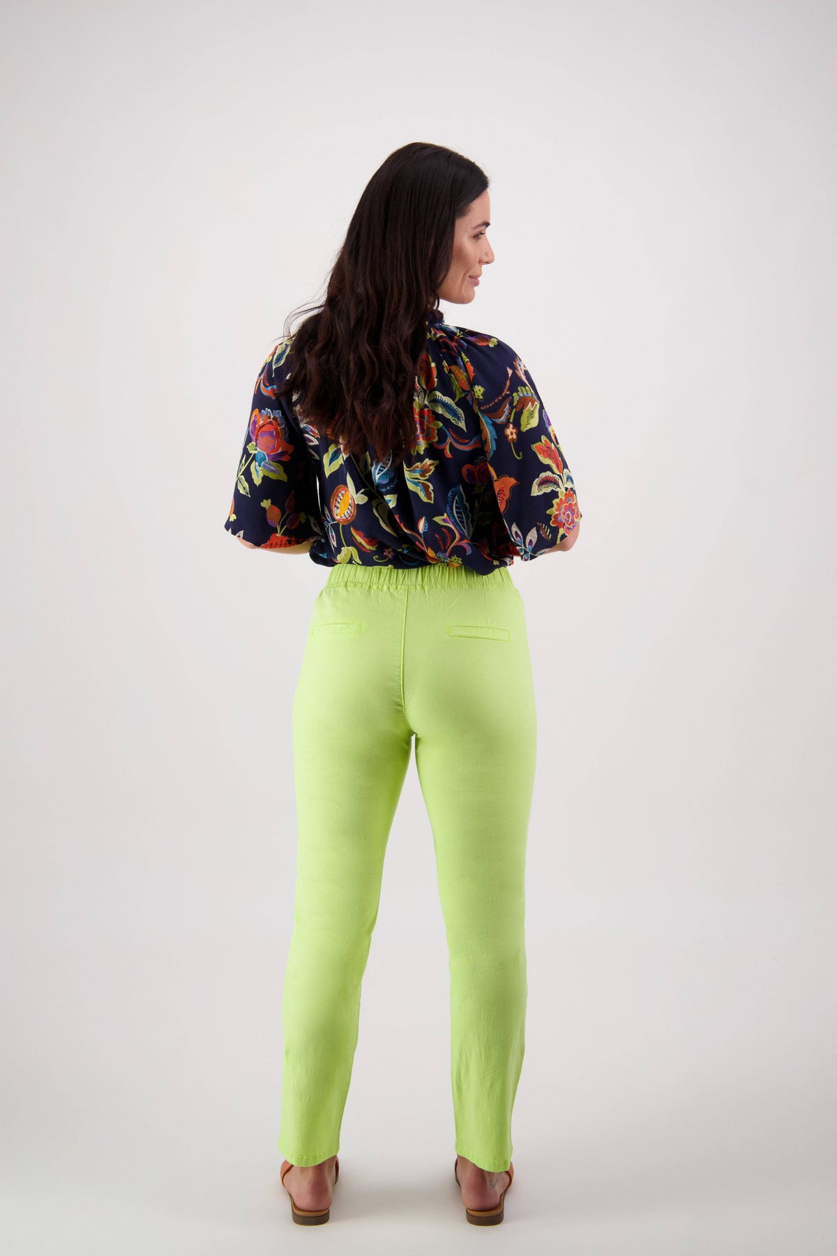 A woman stands facing away against a plain white background, wearing a dark floral blouse paired with Vassalli’s Leonie Pant in Lime—light green ankle-grazer pants featuring an elastic waist and slim leg—and sandals.