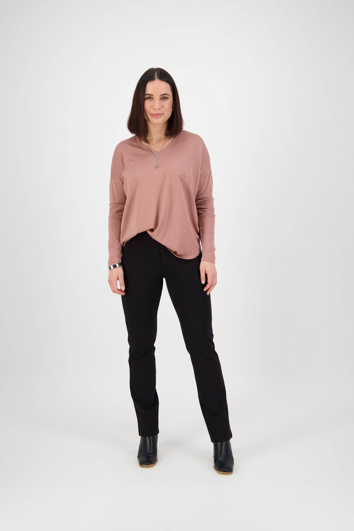 A woman with shoulder-length brown hair faces forward against a white background, wearing a loose pink long-sleeve top, black boots, and Vassalli Slim Leg Ponti Pant in black.