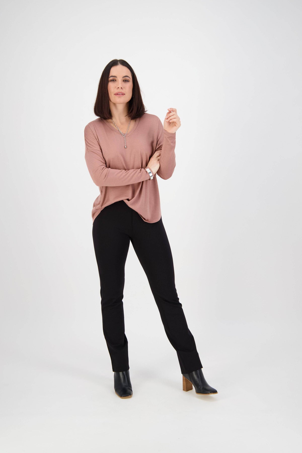A woman with straight dark hair stands against a white background, wearing Vassalli's Slim Leg Ponti Pant in black and a pink long-sleeve top. She looks at the camera with a neutral expression, one hand raised.