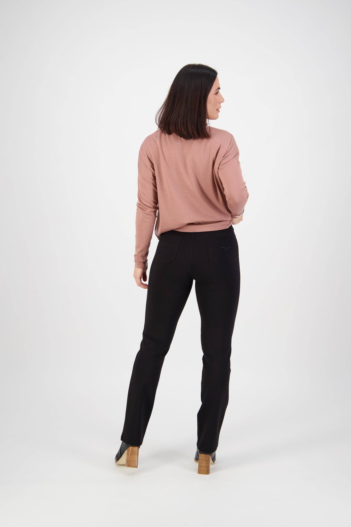 A woman with shoulder-length dark hair wears a pink long-sleeve top and Vassalli Slim Leg Ponti Pant - Black, made from stretch fabric, standing with her back to the camera against a plain white background.