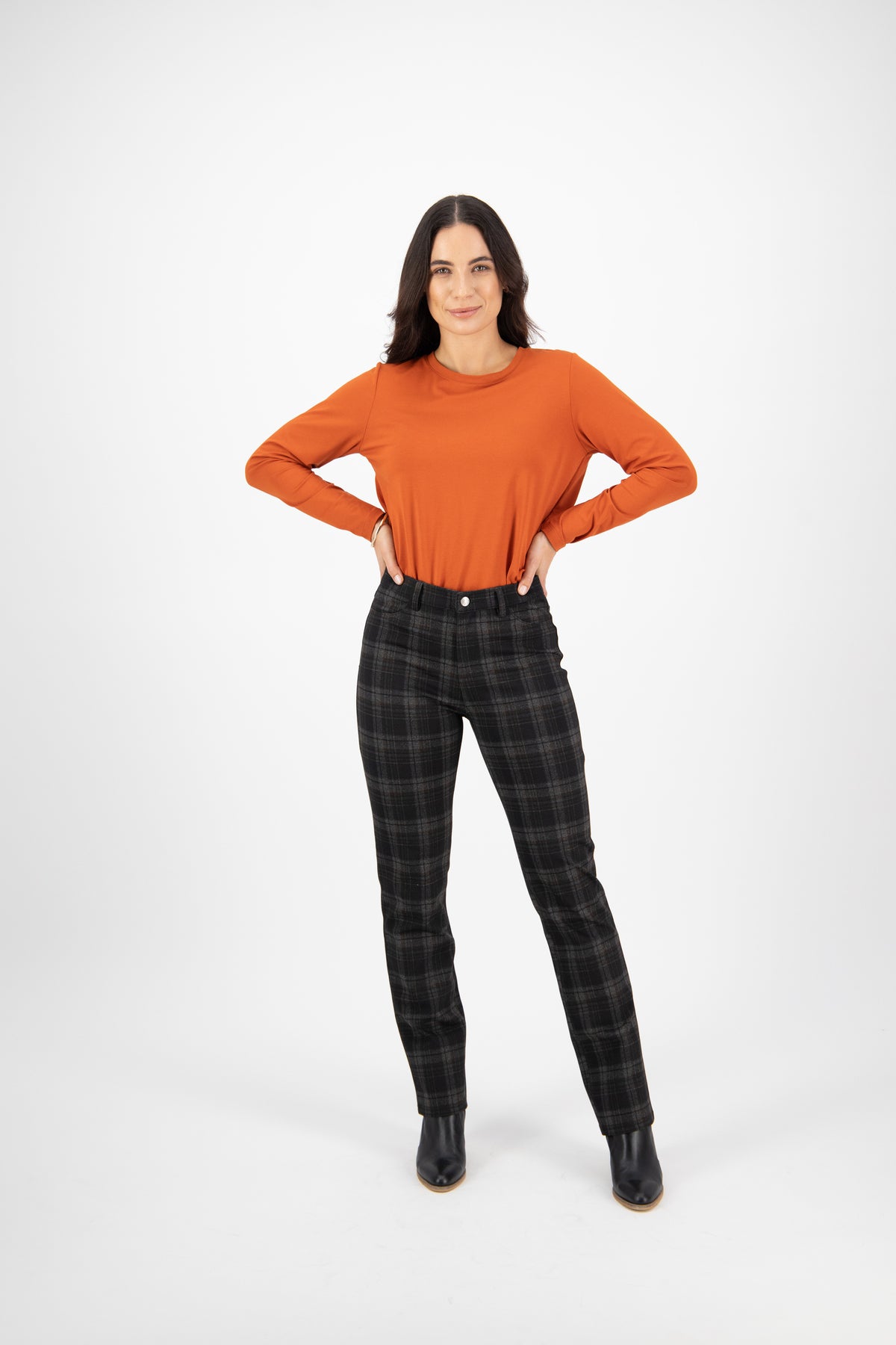 A woman stands confidently against a white background, wearing a long-sleeved orange top, Vassalli Slim Leg Ponti Pant - York in black and gray plaid, and black ankle boots, with her hands on her hips and a slight smile.