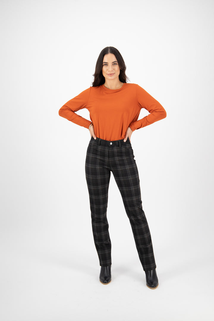 A woman stands confidently against a white background, wearing a long-sleeved orange top, Vassalli Slim Leg Ponti Pant - York in black and gray plaid, and black ankle boots, with her hands on her hips and a slight smile.