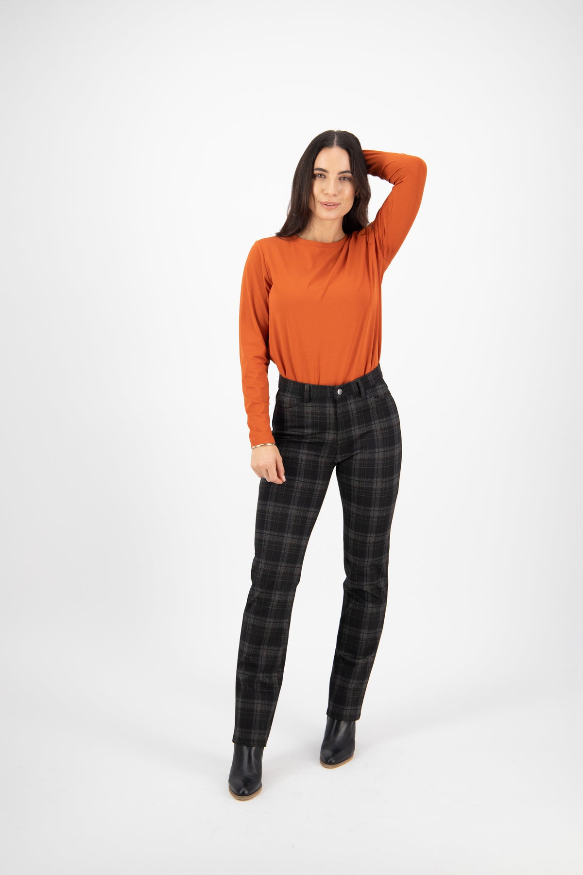 A woman with long dark hair stands on a white background, wearing an orange long-sleeve top, Vassalli Slim Leg Ponti Pant - York in black, and black shoes. She has one hand resting on her head and is smiling slightly.
