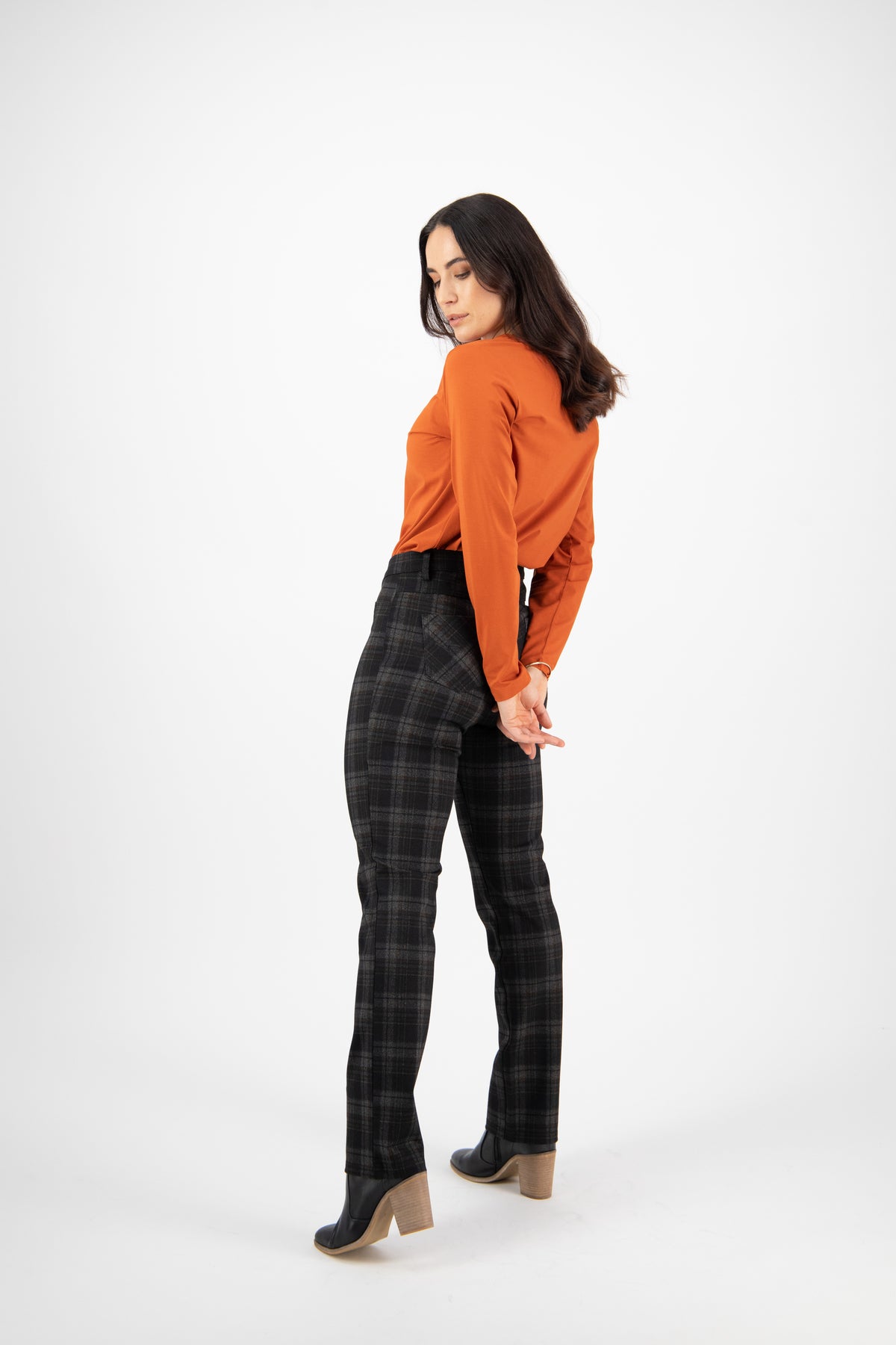 A woman stands against a white background, looking down over her shoulder. She wears an orange long-sleeve top, Vassalli Slim Leg Ponti Pant - York in dark plaid, and black ankle boots with wooden heels, hands clasped behind her back.