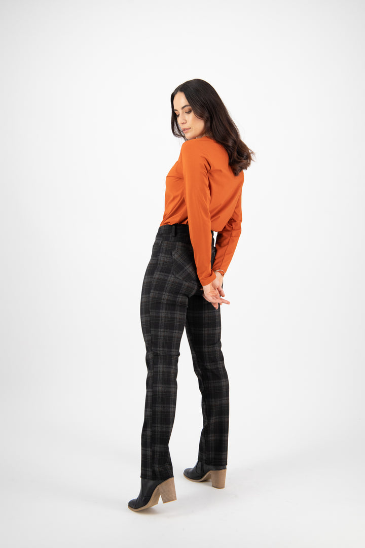 A woman stands against a white background, looking down over her shoulder. She wears an orange long-sleeve top, Vassalli Slim Leg Ponti Pant - York in dark plaid, and black ankle boots with wooden heels, hands clasped behind her back.