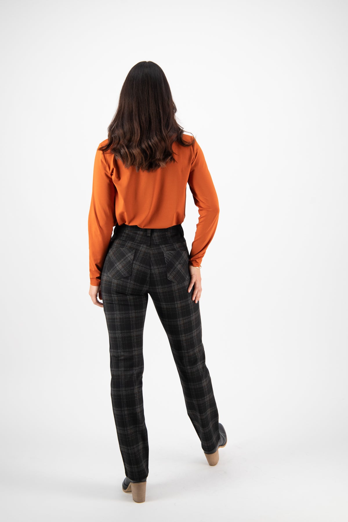 A woman with long brown hair faces away, wearing an orange long-sleeve shirt and Vassalli Slim Leg Ponti Pant - York with heeled shoes, against a plain white background.