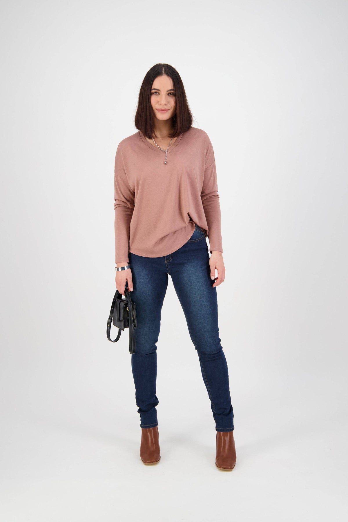 A woman faces forward against a plain white background, wearing a pink long-sleeve top, Vassalli Skinny Leg Jean - Deep Blue, brown ankle boots, and holding a black handbag.