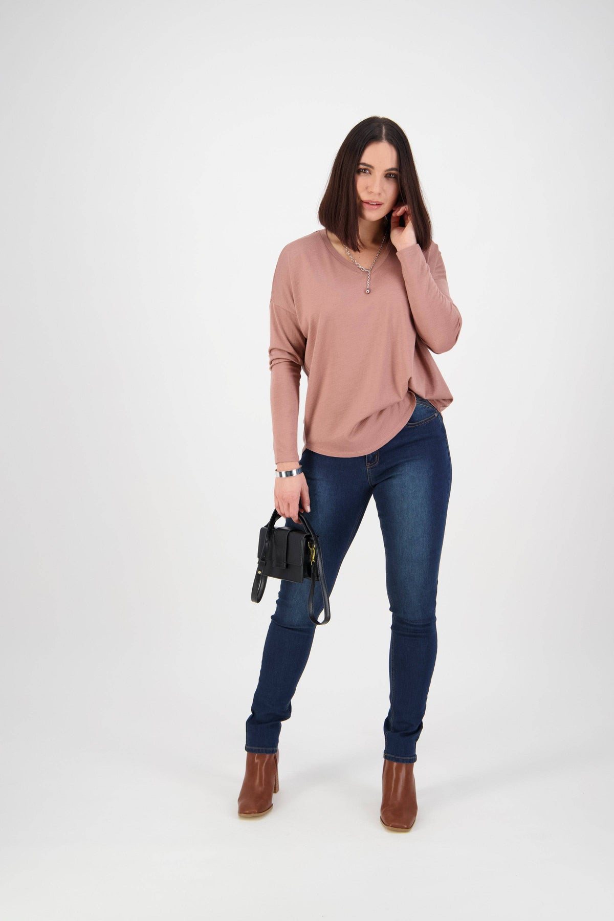 A woman with straight dark hair wears Vassalli's Skinny Leg Jean in Deep Blue, a mauve long-sleeve top, brown ankle boots, and holds a small black handbag. She stands against a plain white background, touching her hair and smiling.