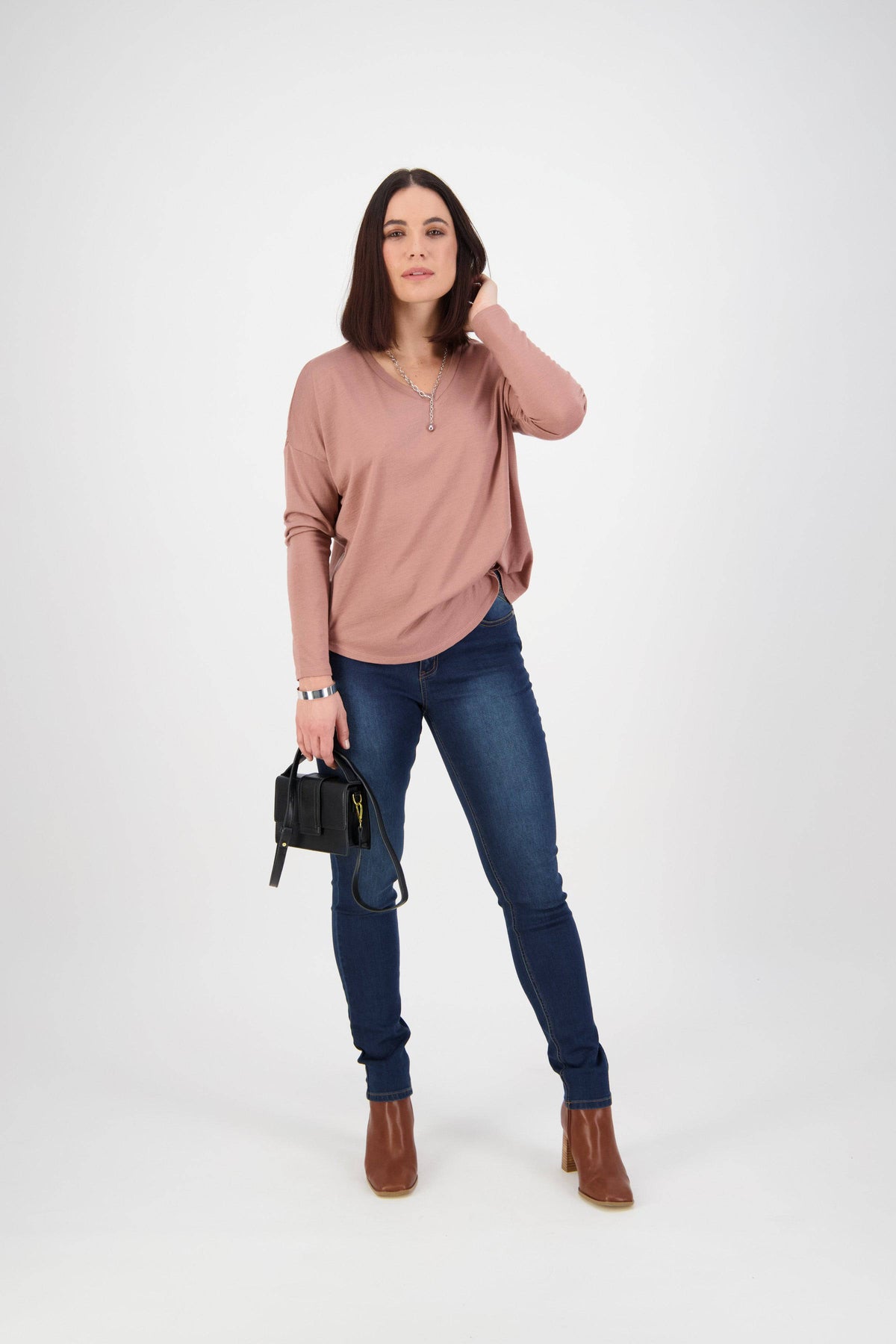 A woman with shoulder-length dark hair wears a pink long-sleeve top, Vassalli Skinny Leg Jean - Deep Blue, and brown ankle boots. She holds a small black purse, standing against a white background with one hand touching her hair.