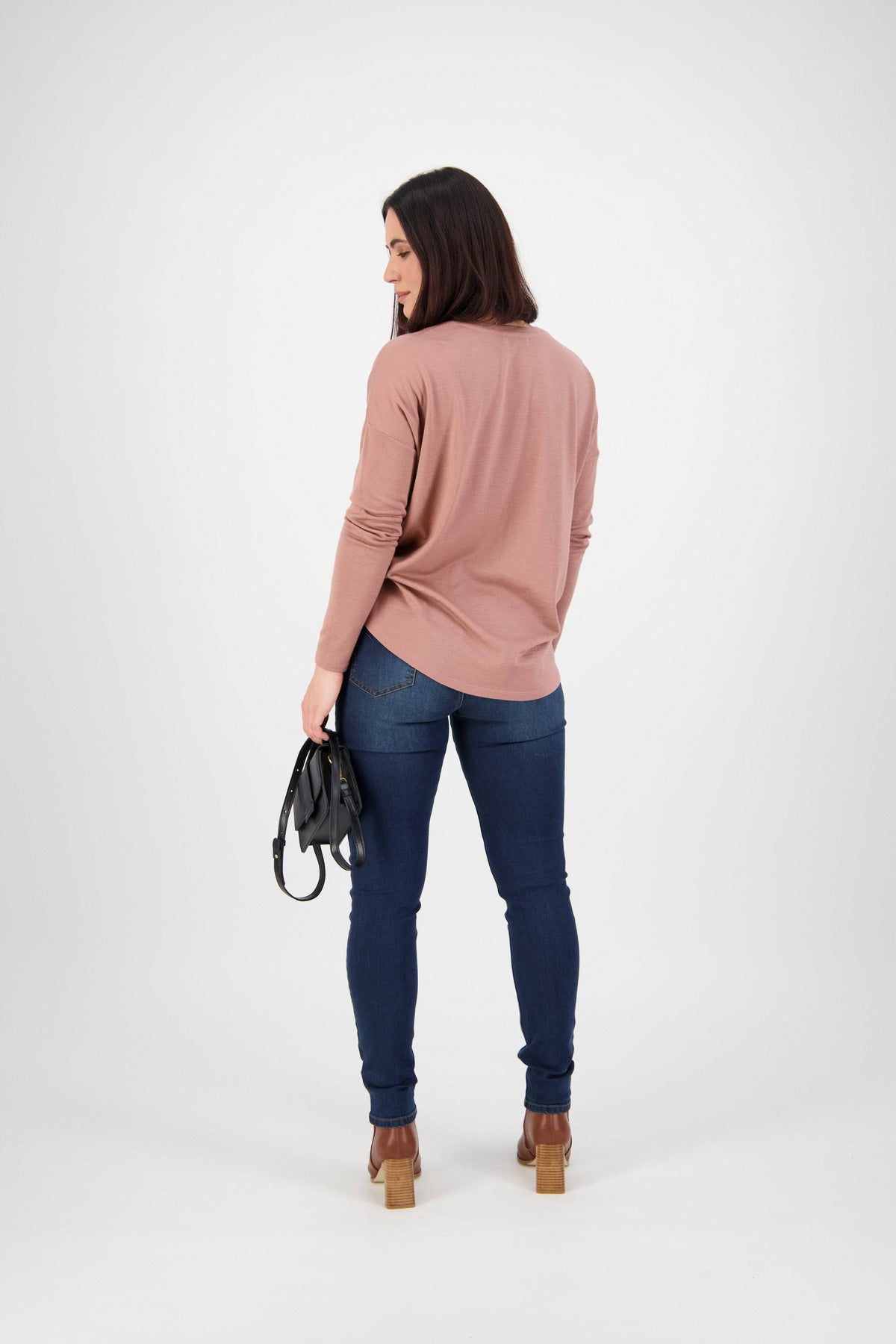 A woman with shoulder-length dark hair, in a long-sleeve pink top and Vassalli Skinny Leg Jean - Deep Blue, brown heeled boots, holds a small black handbag in her left hand while facing away against a plain white background.