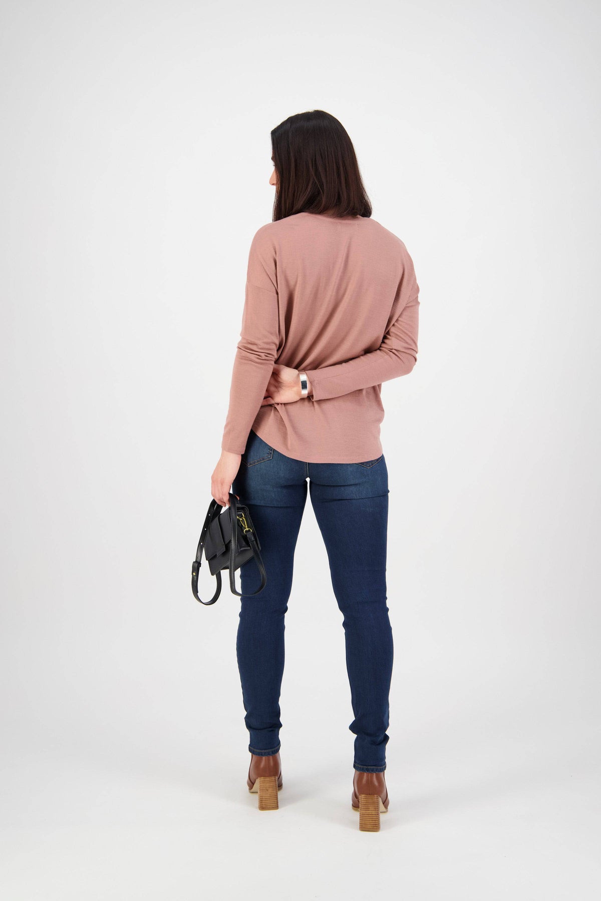 A woman with shoulder-length dark hair stands facing away, wearing a light brown long-sleeve top, Vassalli Skinny Leg Jean - Deep Blue, and heeled shoes. She holds a small black handbag against a plain white background.