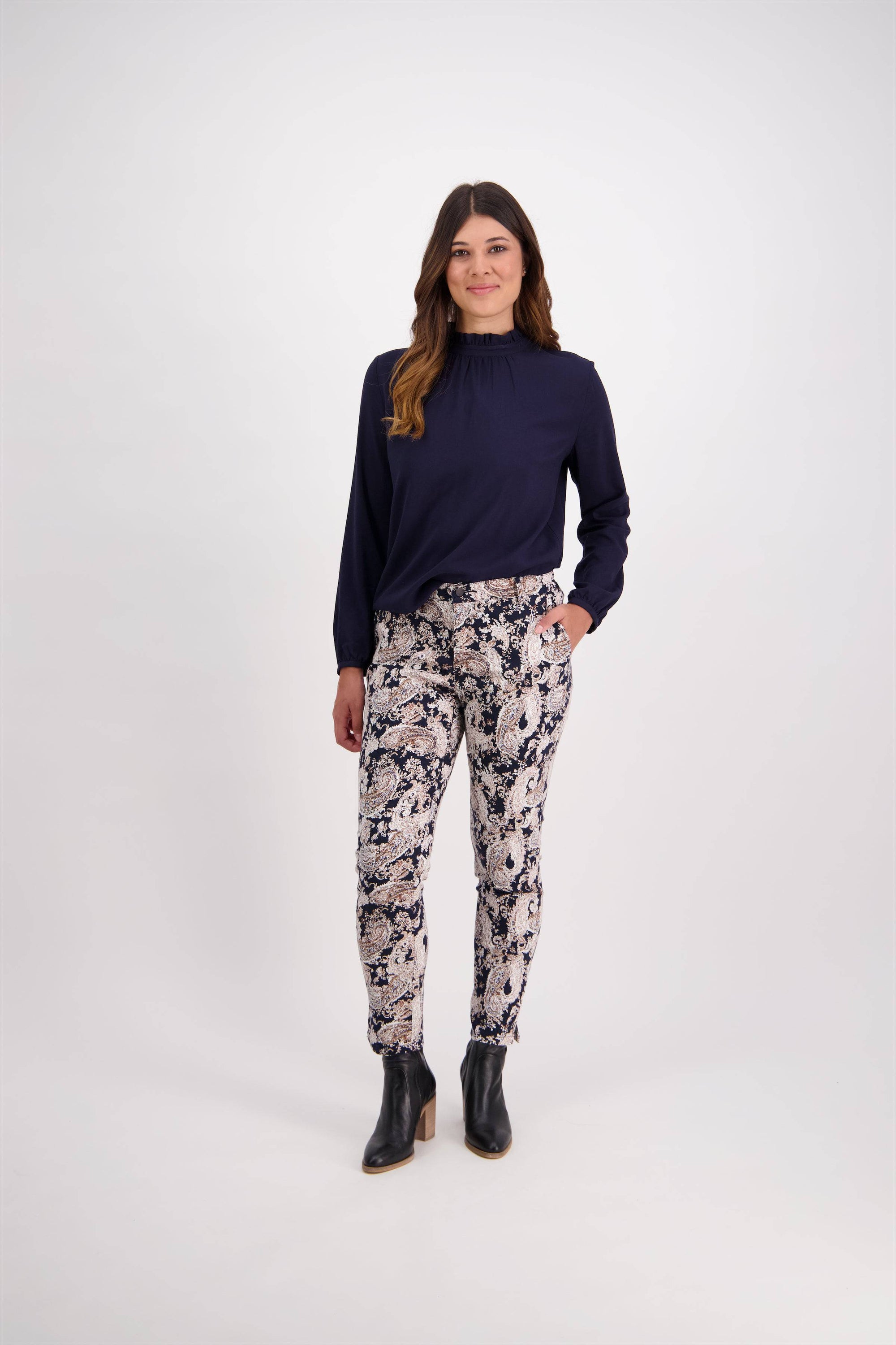 A woman with long, wavy brown hair and a soft smile stands against a white background, wearing a navy long-sleeve blouse, Vassalli Ankle Grazer Pant - Antique with stretch fabric, and black ankle boots.