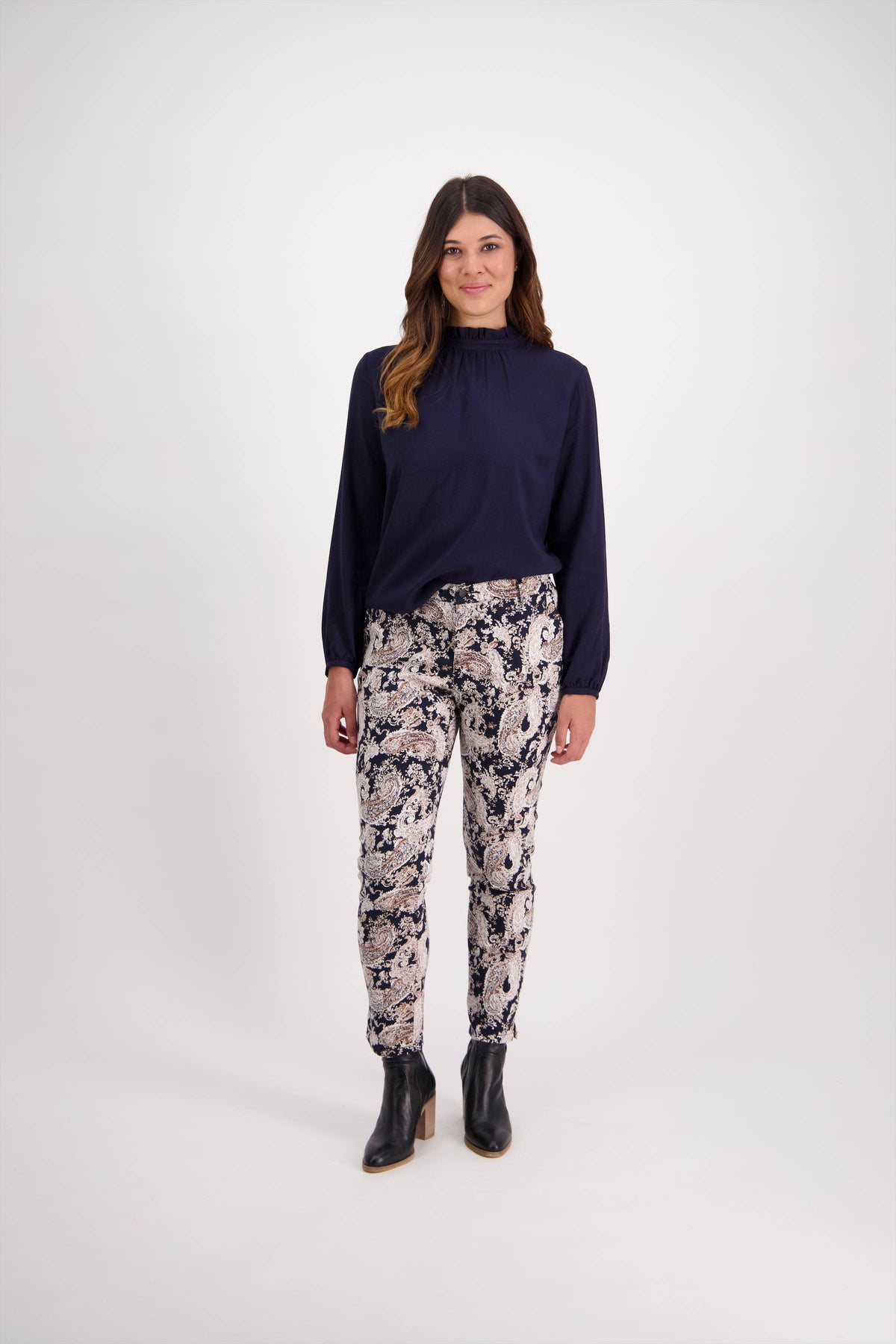 A woman with wavy brown hair smiles softly at the camera, standing against a white background in a long-sleeved navy blouse, Vassalli Ankle Grazer Pant - Antique, and black ankle boots.